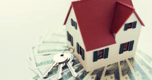 Model house set on top of a fanned out stack of cash with a set of keys to the side.