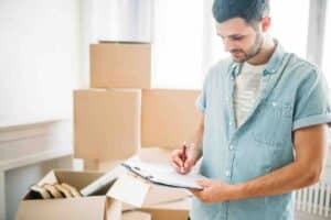 Man in a denim shirt by moving boxes making a list to organize for cross-country moves.