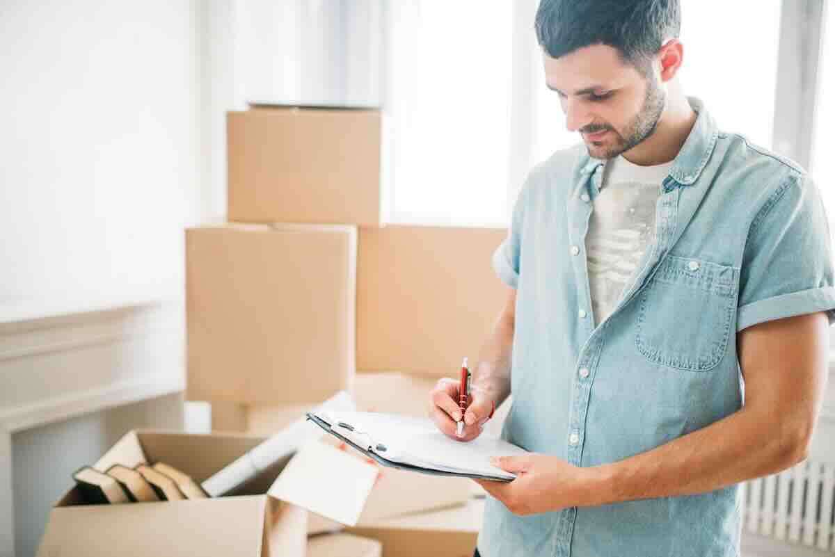 Man in a denim shirt by moving boxes making a list to organize for cross-country moves.