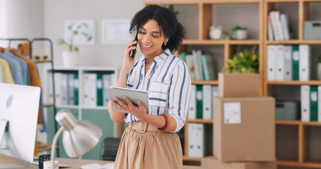 Woman in a striped shirt talking on a phone about moving company prices while researching moving company quotes on a tablet.